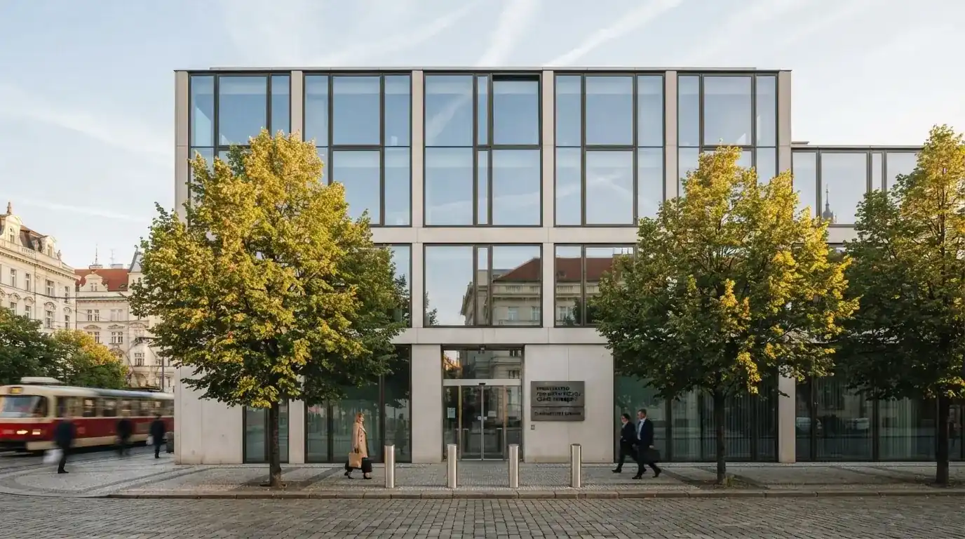 Fasáda moderní administrativní budovy s skleněnými okny odráží ranní oblohu nad rušnou pražskou ulicí.